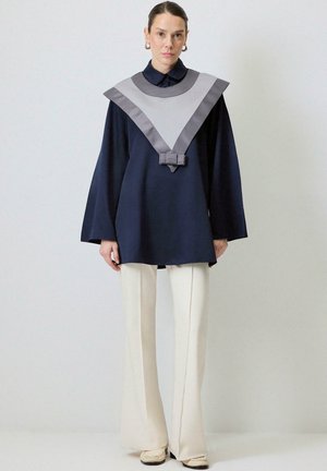 Tunica navy con colletto in satin grigio, caratterizzata da un design a forma di V e un dettaglio a fiocco in vita, abbinata a pantaloni crema a gamba svasata.