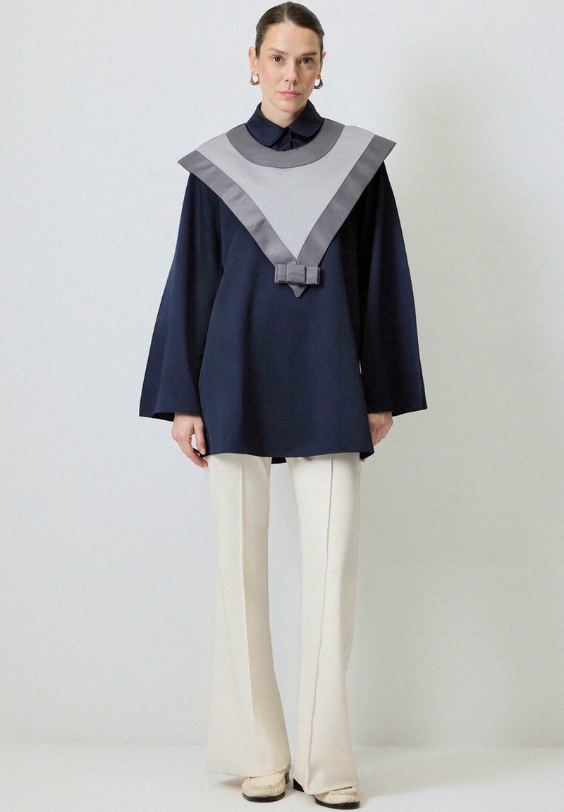 Tunic albastru marin cu guler din satin gri, având un design în formă de V și detalii cu funda la talie, asortată cu pantaloni evazați de culoare crem.