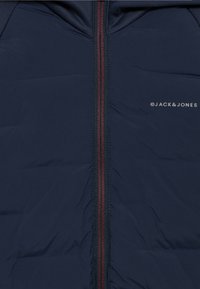 Σκούρο μπλε μπουφάν τύπου puffer με κόκκινο φερμουάρ. Διαθέτει κουκούλα και λογότυπο τυπωμένο στη δεξιά πλευρά. Ομαλή υφή με ραφή τύπου καπιτονέ.