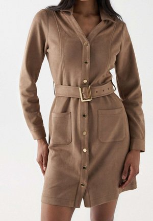 Femme portant une robe-chemise beige ceinturée à manches longues, avec des poches avant et des boutons-pression dorés sur le devant.