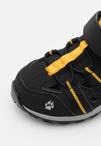 Musta urheilukenkä keltaisilla yksityiskohdilla, säätöremmeillä, teksturoitulla päällä ja harmaalla ulkopohjalla, jossa on paw print -logo.