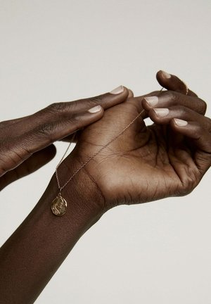 Collar con colgante de oro y una delicada cadena, sostenido entre dos manos. El colgante presenta un diseño ovalado texturizado sobre un tono de piel oscuro.