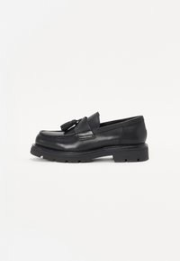 Mocasines de cuero negro con detalles de borlas, punta cuadrada y suela de goma gruesa. Textura suave con costuras tonales y acentos mínimos.