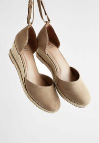 Espadrilles compensées en daim beige avec brides autour de la cheville et semelles plateforme en jute tressé, suspendues sur un fond blanc.