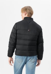 Geacă puffer neagră cu quilt-ing orizontal, guler înalt și un detaliu mic cu un logo tricolor pe spate. Materialul pare neted și izolat.