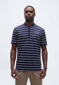 Άνδρας που φοράει κοντομάνικο πουκάμισο Calvin Klein με ρίγες σε ναυτικό μπλε και λευκό χρώμα, με φερμουάρ στον γιακά, στέκεται μπροστά σε απλό φωτεινό φόντο.