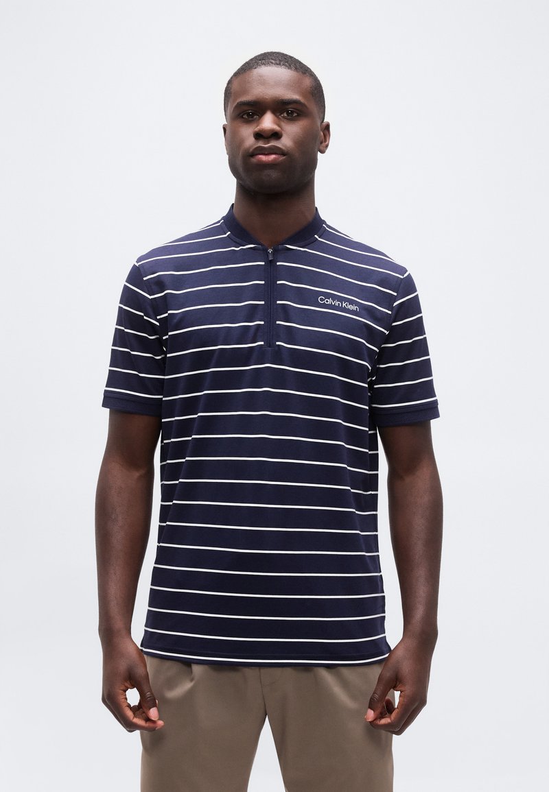 Άνδρας που φοράει κοντομάνικο πουκάμισο Calvin Klein με ρίγες σε ναυτικό μπλε και λευκό χρώμα, με φερμουάρ στον γιακά, στέκεται μπροστά σε απλό φωτεινό φόντο.