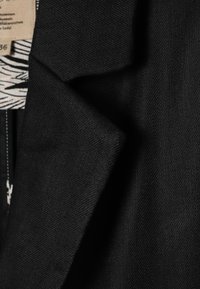 Gros plan sur le revers d'un blazer noir sur mesure avec une texture de tissu visible et une vue partielle d'un vêtement intérieur avec une étiquette à motif.