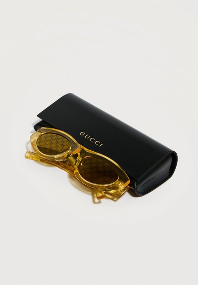 Κίτρινα διαφανή γυαλιά ηλίου Gucci με φακούς διακοσμημένους με σχέδιο που ακουμπούν σε μαύρη θήκη γυαλιών με το λογότυπο Gucci.