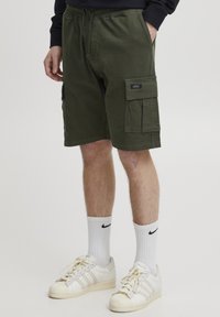Olivenfargede cargo-shorts laget av bomull, med snor i livet og sidelommer. Brukes med hvite Adidas-sneakers og ankelsokker.