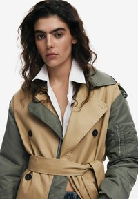 Khaki en olijfgroene trenchcoat met een ceintuur in de taille, grote revers, zwarte knopen en ritszakken op de pols. Gladde en gestructureerde stof.