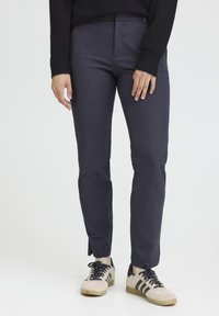 Navy blue tailored trousers made of smooth fabric, featuring a slim fit and side slits at the hem, paired with beige and black sneakers.