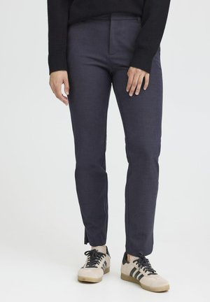 Navy blue tailored trousers made of smooth fabric, featuring a slim fit and side slits at the hem, paired with beige and black sneakers.