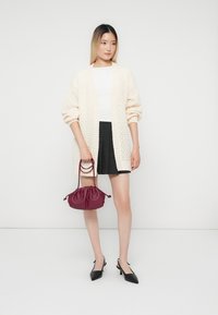 Femme portant un cardigan en tricot crème, un haut blanc, une jupe noire, des escarpins noirs à talon ouvert, tenant un sac à main bordeaux devant un fond blanc uni.