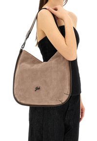 Borsa a tracolla in suede beige con forma curva, rifiniture scure a contrasto e una tracolla. Presenta un logo sulla parte anteriore e cuciture ben visibili.