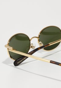 Gafas de sol redondas con lentes verdes, marco de metal dorado, templos adornados y brazos con patrón de carey marrón. Nombre de la marca en el brazo.