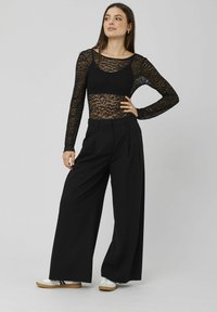 Haut à manches longues en dentelle noire superposé sur un bralette noir, associé à un pantalon noir taille haute à jambe large et des baskets blanches avec des semelles brunes.
