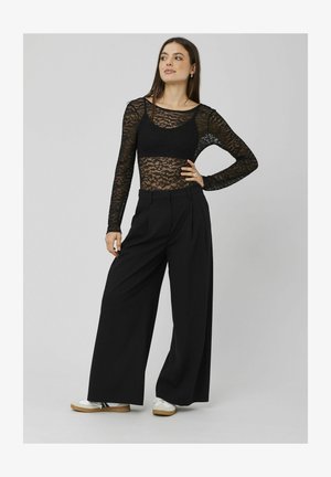 Haut à manches longues en dentelle noire superposé sur un bralette noir, associé à un pantalon noir taille haute à jambe large et des baskets blanches avec des semelles brunes.