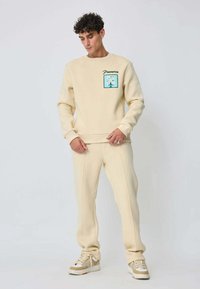 Ensemble de sweatshirt et de pantalon de jogging beige en matière douce, doté d'un motif floral sur la poitrine et de coutures texturées, associé à des baskets.