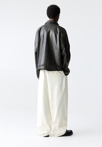 Zwarte leren jassen met kraag en een relaxte pasvorm, gecombineerd met oversized witte broeken en zwarte schoenen, tegen een effen witte achtergrond.