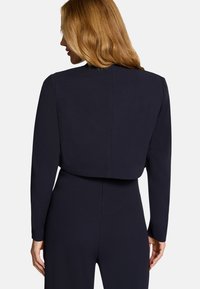 Femme aux longs cheveux blonds portant une combinaison noire ajustée à manches longues, vue de dos sur un fond blanc.