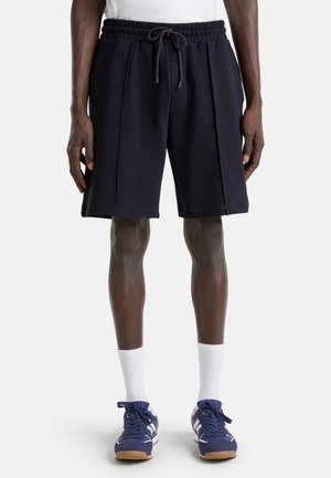 Mann trägt schwarze Shorts mit Kordelzug, weiße Socken und blaue Turnschuhe und steht vor einem einfarbigen Hintergrund.