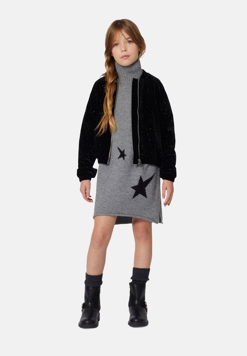 Zwarte fluwelen bomberjack over een grijs gebreid jurkje met zwarte sterpatronen. Het model draagt zwarte enkellaarsjes met gebreide manchet.