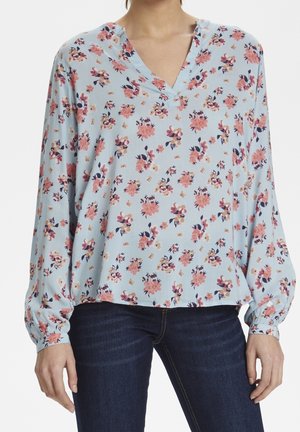 Femme portant un chemisier bleu clair à manches longues avec un motif floral rose et un jean bleu foncé, debout devant un fond uni.