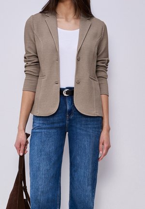 Kvinde iført beige tekstureret blazer over hvid skjorte og blå jeans, der holder en brun ruskindstaske ved siden af sig.