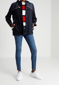 Person klädd i marinblå jacka med fransar, randig tröja i rött, vitt och blått, blå smala jeans och vita sneakers, som står framför en neutral bakgrund.