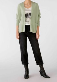Lys grøn strikket cardigan med V-udskæring, to lommer og en enkelt knaplukning, parret med højtaljede sorte jeans og sorte støvler.