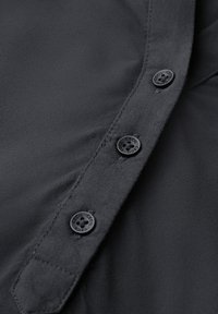 Camisa negra con una textura suave, que presenta un frente de botones con cuatro botones circulares negros y un detalle de cuello tipo punta.