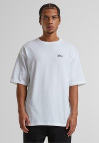 Hvid bomulds-T-shirt med løs pasform, korte ærmer og detalje af sort logo på venstre bryst. Glat tekstur og simpelt design.