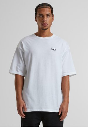 Camiseta de algodón blanca con corte holgado, mangas cortas y un detalle de logo negro en el lado izquierdo del pecho. Textura suave y diseño sencillo.
