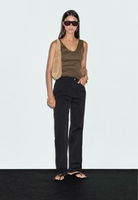 Een vrouw staat zelfverzekerd in een bruine tanktop, zwarte losse broek en sandalen, terwijl ze een beige tas vasthoudt, tegen een lichte achtergrond.