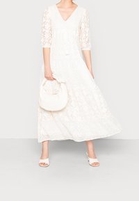 Abito maxi bianco in pizzo con scollo a V, maniche a tre quarti e vita con laccetto. La modella tiene in mano una piccola borsa bianca con dettaglio a nodo.