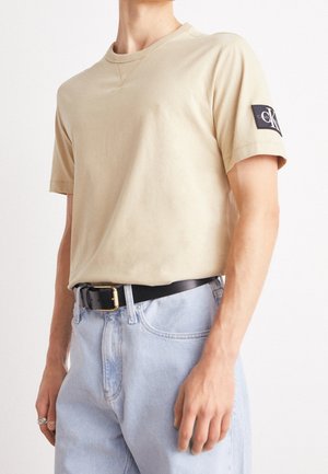 Personne portant un t-shirt Calvin Klein beige à manches courtes, un jean bleu clair, une ceinture noire avec une boucle dorée, et une bague argentée à la main gauche.