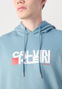 Svetlomodrá mikina vyrobená z jemného materiálu. Obsahuje biele logo s červeným akcentom. Nastaviteľné šnúrky so striebornými koncami. Štandardný strih.