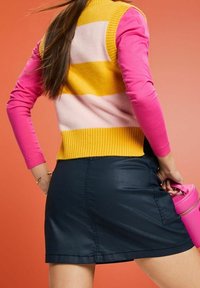 Dzianinowy bezrękawnik w żółte, różowe i białe paski, zestawiony z ciemną mini spódnicą i trzymający małą różową torebkę. Tkanina o fakturze.