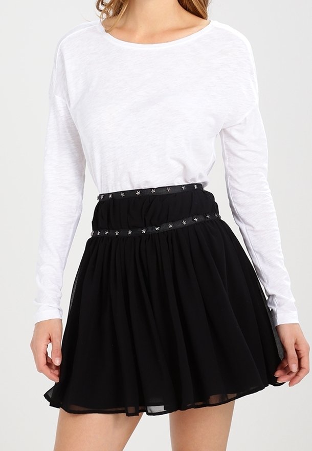 Kvinna klädd i en långärmad vit topp och en svart kjol med ett stjärnmönstrat midjeband, stående mot en neutral bakgrund.