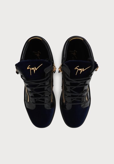Ζευγάρι μαύρα ψηλά sneakers με χρυσά φερμουάρ και λογότυπο στη γλώσσα, σε θέαση από πάνω σε απλό φόντο.
