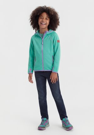 Usmievajúce sa dieťa stojace s uvoľnenými rukami, oblečené v tyrkysovej bunde s purpurovým lemovaním, tmavých džínsoch a ladiacich tyrkysovo-purpurových teniskách.