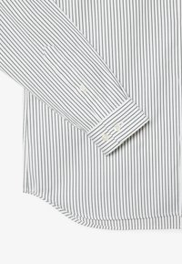 Rokav bele srajce z belimi tankimi črnimi navpičnimi črtami, z zapetim manšetnikom in ukrivljenim robom, ki je viden na ravni površini.