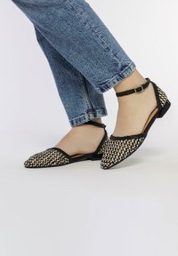 Bottines plates tissées noires et blanches avec un bout pointu, une bride de cheville et un motif texturé sur le dessus, associées à un jeans bleu court.