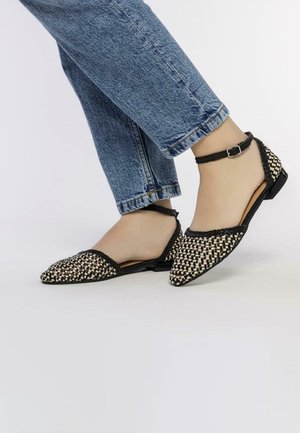 Bottines plates tissées noires et blanches avec un bout pointu, une bride de cheville et un motif texturé sur le dessus, associées à un jeans bleu court.
