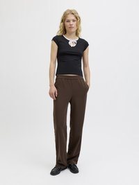 Top negro ajustado con cuello blanco y detalle de lazo, combinado con pantalones marrones sueltos. La modelo está de pie en un fondo neutro.