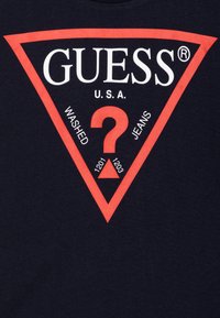 Sötétkék póló, nagy piros háromszög grafikával. A szöveg tartalmazza a "GUESS," "U.S.A.," és "WASHED JEANS" feliratokat, a közepén egy kérdőjellel.