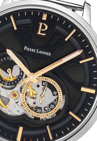 Cadran noir avec accents dorés, affichant des mouvements mécaniques. Présente un logo de marque proéminent et un design de cadran unique avec des découpes.