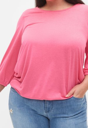 Femme portant un haut rose ample à manches trois-quarts avec un jean bleu clair, la main droite dans la poche.