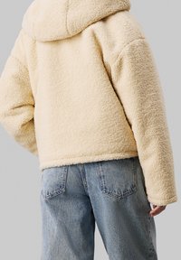 Kremowy, pluszowy, kapturowy płaszcz o teksturowanej powierzchni. Luźny krój, skrócona długość, zestawiony z jasnoniebieskimi dżinsami.